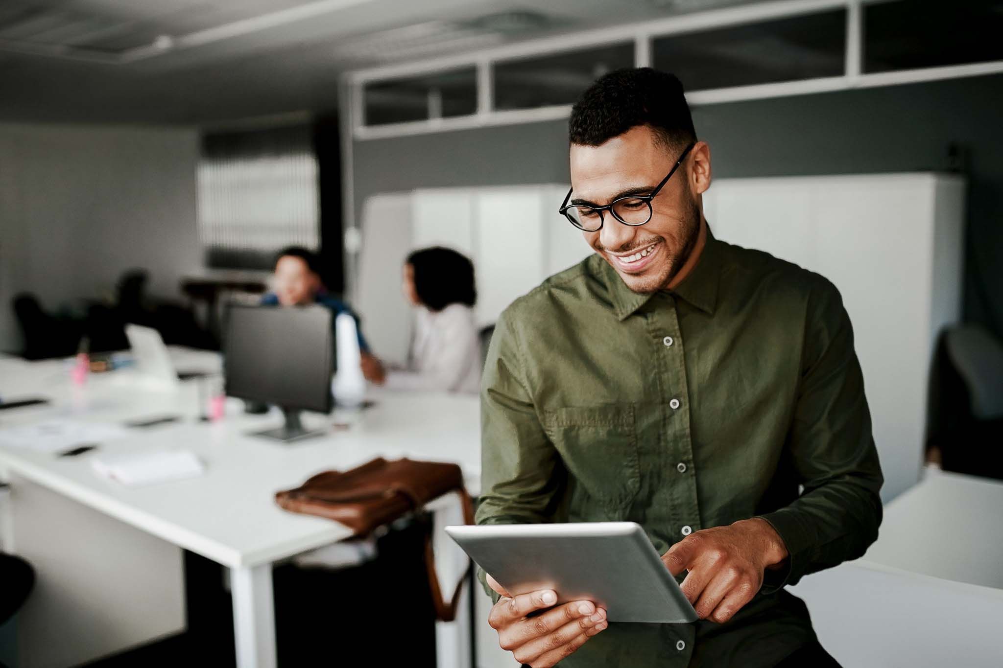 Cheerful young businessman using digital tablet smiling while his colleagues working at the background